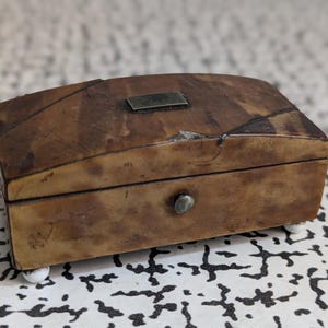 May include: A rectangular wooden box with a curved lid, featuring a mottled brown finish, a small metal rectangular plate on top, a single metal knob on the front, and four small white spherical feet supporting the base of the container.