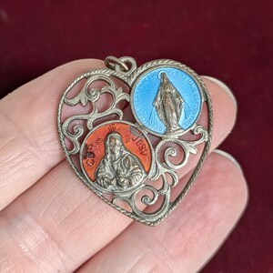 May include: Vintage silver-toned heart-shaped pendant with two circular enamel insets. One inset features a blue image of the Virgin Mary, the other an image of Jesus. The pendant has ornate filigree detailing and a small loop for a chain.