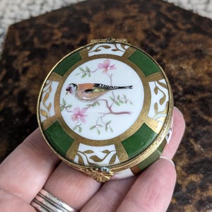 May include: A small, round, porcelain trinket box with a gold and green design. The lid features a hand-painted image of a bird perched on a branch with pink flowers.