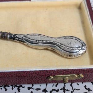 Puede incluir: Un mango de utensilio de metal plateado con un extremo alargado y bulboso que presenta un patrón decorativo de borde con cuentas. El mango se estrecha hacia un cuello cilíndrico y descansa dentro de un estuche rectangular con interior color crema.