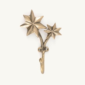 Puede incluir: Un gancho de pared de color latón con un diseño de dos estrellas. Las estrellas son de diferentes tamaños y están conectadas por un tallo curvo. El gancho está diseñado para colgar objetos y tiene una apariencia decorativa clásica.