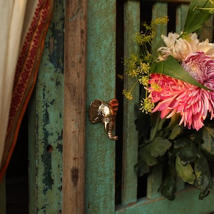 Op de afbeelding: Een gouden deurknop in de vorm van een olifant met een getextureerd oppervlak is gemonteerd op een turquoise houten deur. De deur is verweerd en heeft een rustieke uitstraling. Op de achtergrond is een boeket roze en witte bloemen te zien.