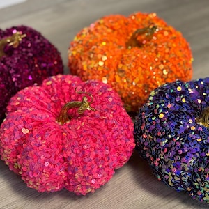 Puede incluir: Cuatro calabazas decorativas cubiertas de lentejuelas. Las calabazas son en tonos de morado, naranja, rosa y azul.