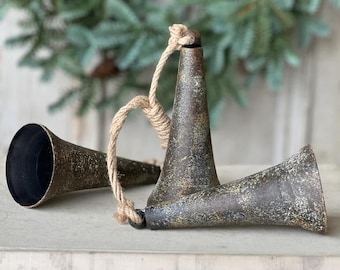 May include: Two antique-style metal bells with a distressed, bronze finish. Each bell has a flared opening and a rope handle. The bells are resting on a stack of vintage books, with a blurred green wreath in the background.