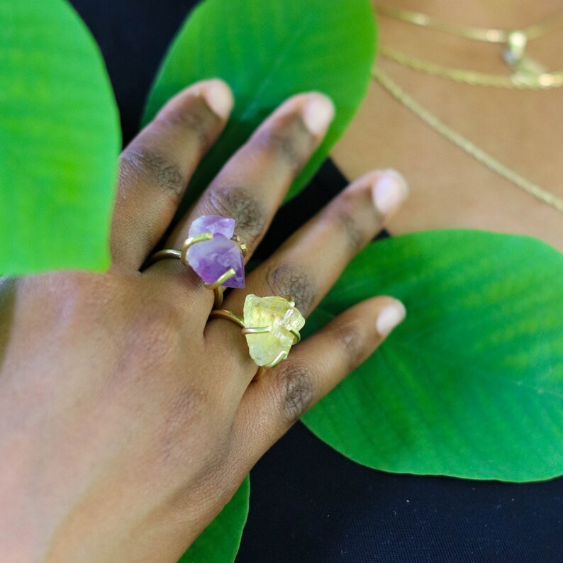 May include: Two gold rings with raw crystal stones. The ring on the left has a purple amethyst stone and the ring on the right has a yellow citrine stone.