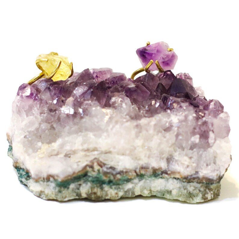 May include: Two rings with amethyst and citrine gemstones set in gold, displayed on a bed of purple amethyst crystals.