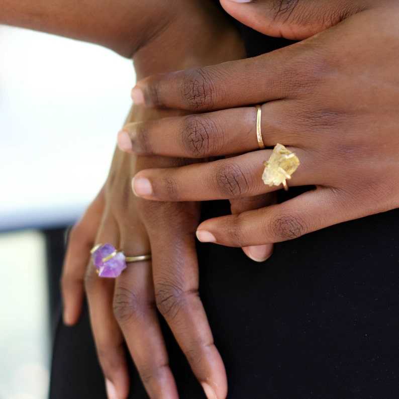 May include: Two gold rings with raw crystal stones. The ring on the left has a purple amethyst stone. The ring on the right has a yellow citrine stone.