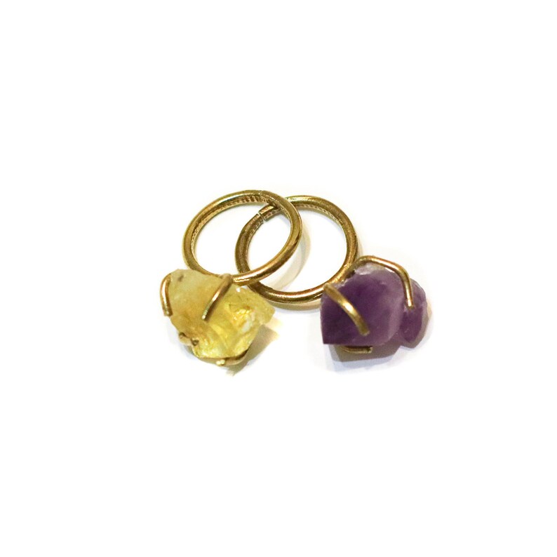 May include: Two gold rings with raw gemstone settings. One ring features a yellow citrine stone and the other a purple amethyst stone.