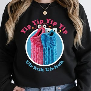 May include: Black sweatshirt featuring a graphic of two fuzzy, sock-like creatures in pink and blue, with googly eyes, inside a white circle. The text "Yip Yip Yip" and "Uh-huh Uh-huh" are in pink and blue, respectively, above and below the creatures.