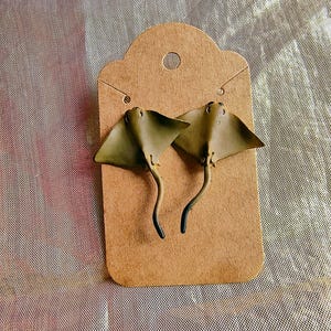 May include: A pair of olive green stingray earrings displayed on a brown earring card. The earrings have a detailed design, with a curved tail and a textured surface. The card has a hole at the top for hanging.