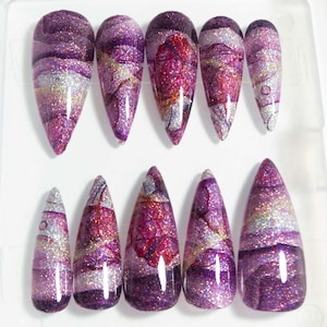 May include: A set of ten artificial nails in a clear plastic container. The nails feature a marbled design with purple, silver, and gold glitter. The nails are long and pointed, with a glossy finish.