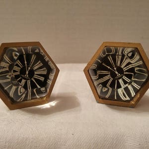 May include: Two hexagonal clock-shaped objects with gold-colored frames. The clock faces are black with white Roman numerals and hands. The clocks are displayed on a white surface.