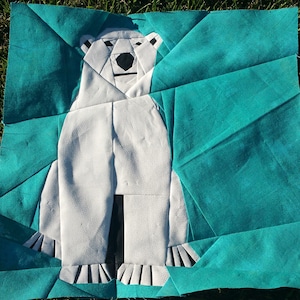 Op de afbeelding: Een witte poolbeer-applique op een turquoise stoffen achtergrond. De beer is gemaakt van stofstukken die aan elkaar zijn genaaid om een geometrisch ontwerp te creëren.