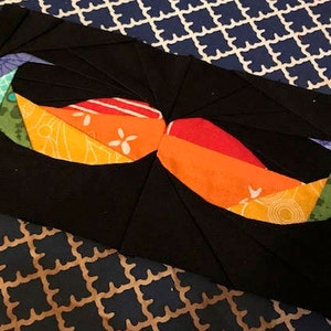 Puede incluir: Un bloque de colcha de tela negra con un diseño geométrico colorido que se asemeja a un par de bigotes. El diseño presenta una variedad de colores, incluyendo naranja, rojo, amarillo, verde, azul y turquesa.