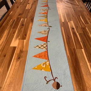 Może przedstawiać: Niebieski pikowany bieżnik stołowy z sznurkiem z materiałowych chorągiewek w różnych jesiennych kolorach i wzorach. Chorągiewki są przyszyte do brązowej gałęzi z brązowym żołędziem na dole.