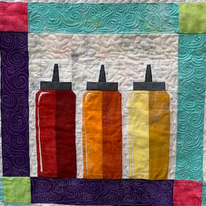 May include: Three colourful glass bottles with black tops, resembling condiment bottles, are arranged in a row on a white background. The bottles are filled with red, orange, and yellow liquids.