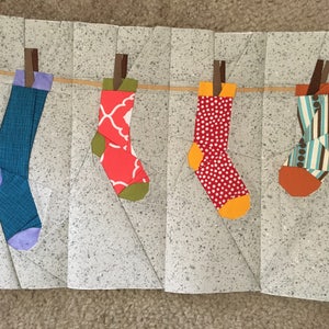 May include: Four colorful socks hanging on a clothesline against a gray background. The socks are a teal blue with purple trim, a red with white design and green trim, a red with white polka dots and orange trim, and a blue and brown striped sock with orange trim.