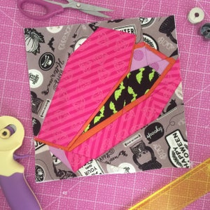 May include: A pink and orange fabric coffin with a green and black bat print lining. The coffin is on a gray fabric background with Halloween-themed text and images.