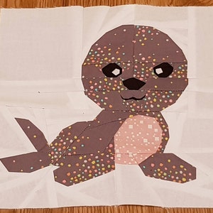 May include: A gray and pink fabric seal applique with a white background. The seal has a speckled pattern and black eyes.