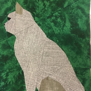 Op de afbeelding: Een grijze kat zittend op een groene achtergrond. De kat is gemaakt van stof en is een patchwork van verschillende stukken. De achtergrond is ook gemaakt van stof en heeft een groen en wit marmerpatroon.