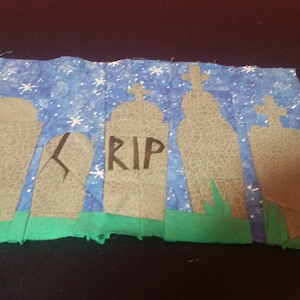 May include: A fabric piece featuring a graveyard scene. Gray tombstones with crosses are set against a blue starry sky. The word "RIP" is visible on one tombstone, with green grass at the bottom. A Halloween or spooky theme.