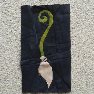 May include: A black fabric patchwork with a green and beige appliqué of a broom with a curled handle.