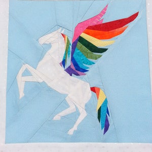 May include: A white paper pieced unicorn with a rainbow wing and tail on a light blue background.