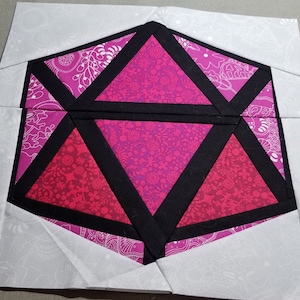 Può includere: Un blocco di trapunta in tessuto rosa e nero con un design geometrico. Il blocco è composto da sei triangoli, con il triangolo centrale nero e i triangoli circostanti rosa.