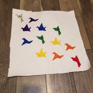 May include: A white fabric square with colorful origami paper birds arranged in a pattern. The birds are in the colors of the rainbow, starting with red and ending with purple.