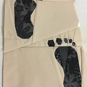 May include: Two beige fabric squares with black and gray floral fabric cutouts in the shape of footprints. The fabric squares are pinned together.
