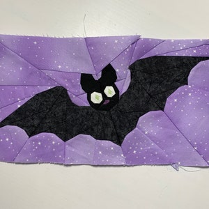 May include: A black bat with green eyes flying against a purple background with white stars.