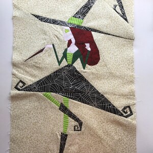 May include: A witch with red hair and a green dress is flying on a broomstick. Her hat is black with a spiderweb pattern. The background is a light beige fabric with a small floral pattern.