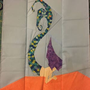 Op de afbeelding: Een zeemeerminstaart gemaakt van stof met een schubbenpatroon in groen, blauw en geel. De staart is opgerold en de bovenkant van de staart is paars. De staart bevindt zich op een lichtblauwe achtergrond met een oranje achtergrond aan de onderkant.