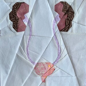 Puede incluir: Un bloque de colcha de tela blanca con un diseño de flor rosa y marrón. La flor tiene un centro rosa y pétalos marrones. El bloque de colcha está hecho de muchas piezas de tela diferentes, que se cosen juntas para crear un patrón geométrico.