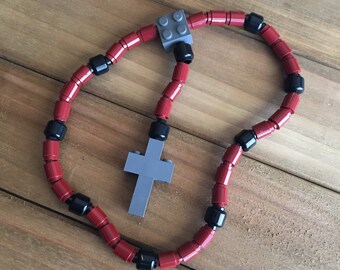 May include: A rosary made of red and black toy blocks, with a gray cross pendant. The beads are cylindrical and connected, forming a loop. The cross and a connecting piece are gray. The rosary is displayed on a wooden surface.