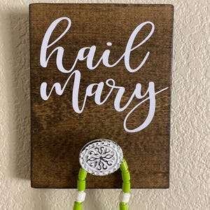 May include: A wooden sign with the words "hail Mary" in white script. The sign has a decorative metal hook with a green and white beaded necklace hanging from it. The wood has a dark brown stain.