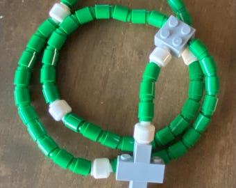 May include: A handmade rosary made from green and white toy building blocks. The rosary features a cross and a series of beads, all constructed from the colorful blocks. The design is a playful take on a traditional religious item.