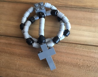 May include: A gray and black rosary made from interlocking toy bricks. The rosary features a cross at the bottom and is composed of gray and black beads. The toy is set against a wooden background.