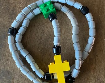 May include: A handmade rosary made from gray and black Lego bricks, with a bright yellow cross and a green Lego brick. The rosary is displayed on a wooden surface.