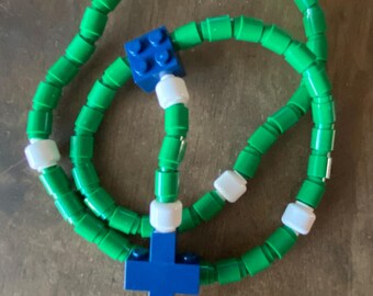 May include: A handmade rosary made of green and white Lego bricks. The rosary features a blue cross and a blue square piece. The beads are green, with white spacers. The rosary is displayed on a wooden surface.