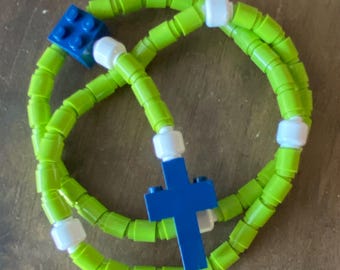May include: A handmade rosary made of green and white plastic beads, with a blue cross and a blue square piece. The rosary is arranged in a circular shape.