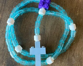 May include: A handmade rosary made of clear blue plastic tubing, white beads, and a light blue cross. A purple Lego brick is used as a decorative element. The rosary is displayed on a wooden surface.