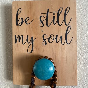 May include: Wooden sign with the words "be still my soul" in black script. A turquoise blue bead is attached to the sign, with a strand of brown wooden beads hanging below.