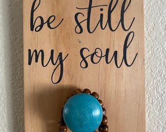 May include: Wooden sign with the words "be still my soul" in black script. A turquoise blue bead is attached to the sign, with a strand of brown wooden beads hanging below.