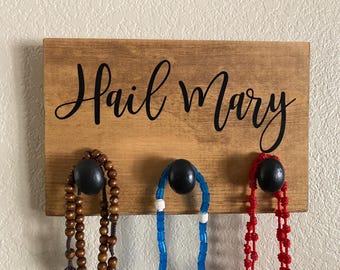 May include: Wooden sign with the words "Hail Mary" in black script. The sign has three black knobs holding rosaries in brown, blue, and red.