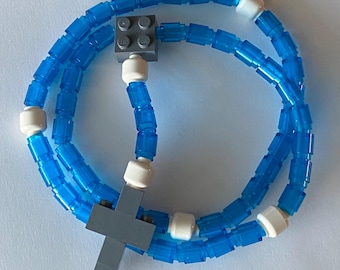May include: A blue and white beaded necklace with a gray cross and a gray square. The necklace is made of small, transparent blue beads and white cylindrical beads. The cross and square are made of gray blocks.
