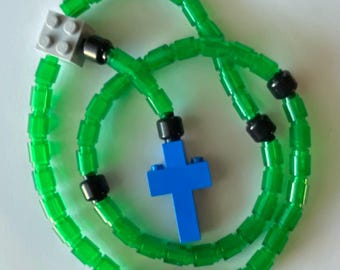 May include: A green rosary made of translucent plastic beads, black connectors, a blue cross, and a gray square block. The beads are strung together to form a loop, with the cross hanging in the center.