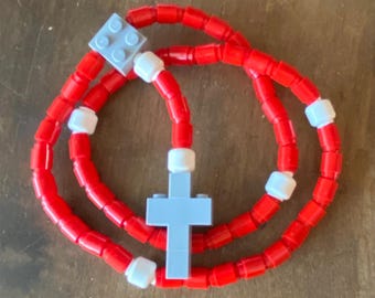 May include: A red rosary made of small, cylindrical beads. The rosary features a light blue cross and a light blue square piece. White beads separate the red beads. The rosary is displayed on a wooden surface.