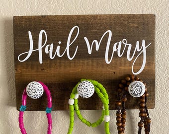 May include: A wooden wall hanging with the words "Hail Mary" in white script. Below the text are three decorative knobs holding necklaces in pink, green, and brown. The knobs have a white, patterned design.