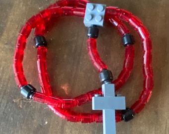 May include: A red beaded necklace with a gray cross pendant and gray square accents. The necklace is made of transparent red beads and black connectors. The cross is constructed from gray building blocks.
